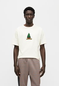 Μπλούζα με κοντά μανίκια σε κρεμ απόχρωση, με σχέδιο πράσινου δέντρου και κείμενο "Converse". Φοριέται με καρό παντελόνι σε απαλούς χρωματισμούς.