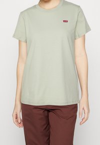 Světlomodré bavlněné tričko s krátkým rukávem, kulatým výstřihem a malým červeným logem Levi's na hrudi. Nosené s hnědými kalhotami.