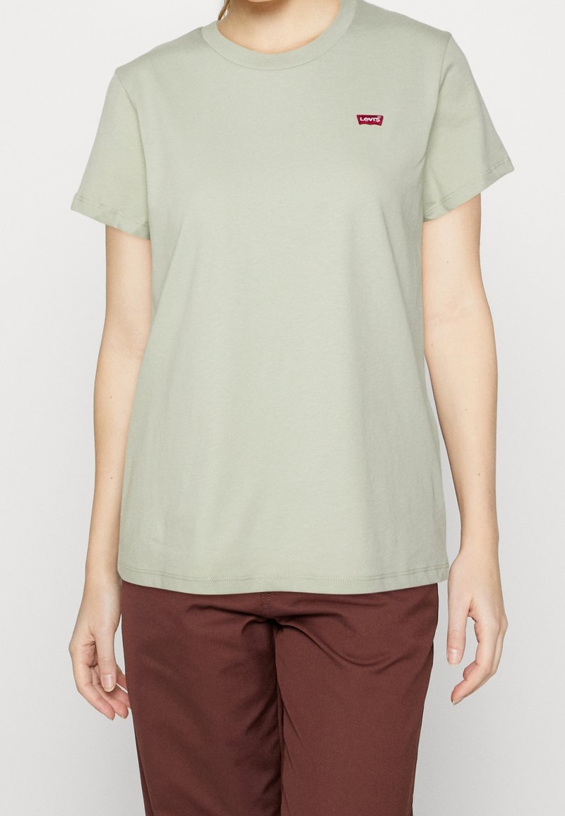 Světlomodré bavlněné tričko s krátkým rukávem, kulatým výstřihem a malým červeným logem Levi's na hrudi. Nosené s hnědými kalhotami.