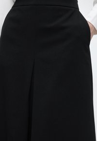 Γυναίκα που φοράει μαύρη φούστα με πιέτα μπροστά, άσπρο πουκάμισο με μακριά μανίκια και τα χέρια της τοποθετημένα στις τσέπες της φούστας.