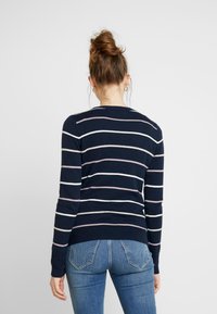 Granatowy sweter w paski z jasnoróżowymi i białymi poziomymi liniami, ściągaczami na mankietach i u dołu, noszony z niebieskimi dżinsami. Miękka dzianinowa faktura.