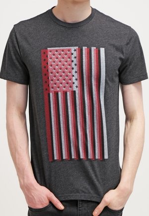 T-shirt en coton gris avec un motif du drapeau américain en rouge, blanc et bleu, comprenant des étoiles, des rayures et une apparence texturée.