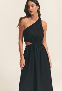 Vestido negro de un solo hombro con cintura elástica, detalle recortado y un lazo decorativo. Tejido suave con una silueta fluida.