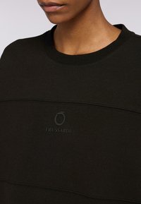 Svart sweatshirt med rund halsringning, prydd med en subtil präglad logotyp och horisontella sömmar. Slät textur, avslappnad passform.