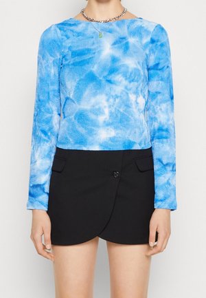 Person iført en langærmet top med blå- og hvide tie-dye mønstre, kombineret med et sort asymmetrisk omslagsnederdel med knap- og lomme detaljer.