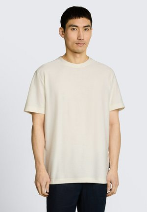 Mężczyzna stojący przodem, ubrany w jednolity beżowy t-shirt z krótkim rękawem i ciemne spodnie na jednolitym jasnoszarym tle.