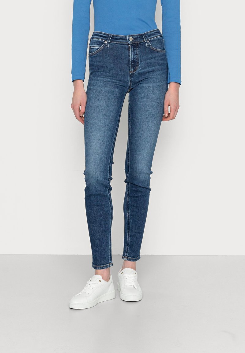 Skinny jeans in medium blue denim, voorzien van vijf zakken, een rits aan de voorkant, metalen knoopsluiting en subtiele stikseldetails. Gekoombineerd met witte sneakers.