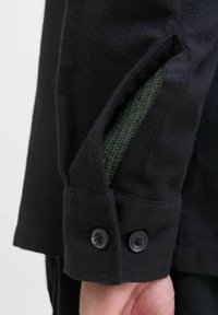 Schwarzer Stoffjackenärmel mit grüner Strickfutter, zwei schwarzen Knöpfen am Bündchen und einem maßgeschneiderten Design mit sichtbaren Nahtdetails.