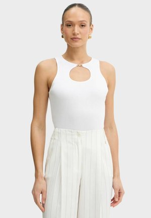 Donna che indossa un top bianco senza maniche con ritaglio frontale e fibbia dorata abbinato a pantaloni bianchi a righe a vita alta su sfondo neutro.