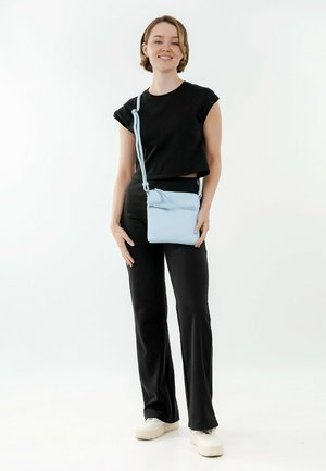 Hellblaues Crossbody-Bag mit strukturiertem Oberflächenmaterial, rechteckiger Form, verstellbarem Riemen und einer Fronttasche mit Reißverschluss, getragen von einer Person in schwarzer Kleidung.
