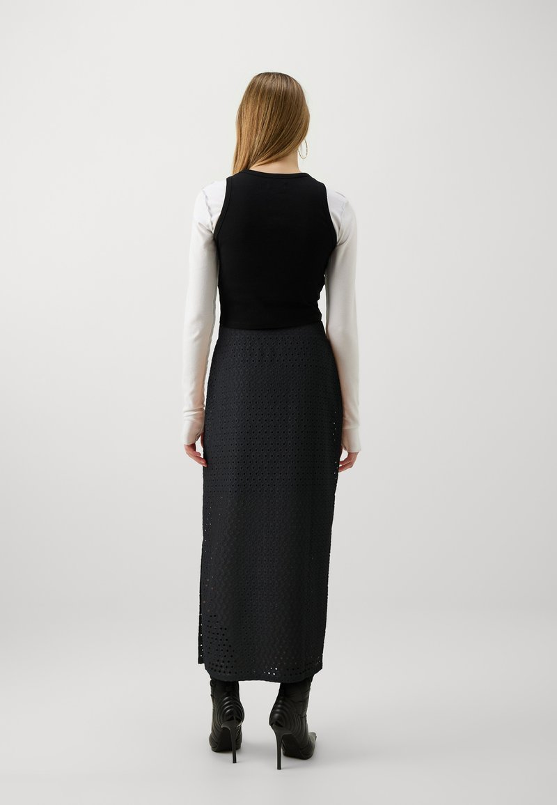 Haut ajusté noir superposé sur une chemise blanche à manches longues, associé à une longue jupe noire perforée. Des bottes à talons noirs complètent le look.
