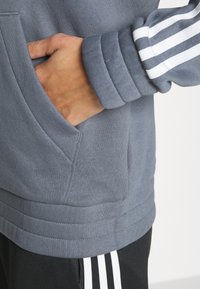 Main dans la poche avant d'un sweat à capuche gris avec des rayures blanches sur la manche, associé à un pantalon noir avec des rayures blanches.