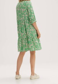 Kobieta w zielonej sukience do kolan w beżowy wzór liści i rękawami 3/4, zestawiona z białymi, sportowymi sneakersami.