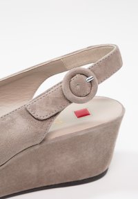 Sandale compensée en daim beige avec une sangle à boucle ronde. Présente une texture lisse et une courbe subtile dans son design. Inclut un accent rouge contrastant.