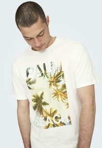 Camiseta de algodón blanca con una gran impresión gráfica de palmeras y el texto "PALM BEACH" en letras grandes. Mangas cortas, cuello redondo.