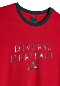 Czerwona koszulka z granatowym dekoltem. Posiada błyszczące srebrne napisy "RÓŻNORODNE DZIEDZICTWO EST. 1993 Oryginalny Klub." Wykonana z miękkiej bawełny.
