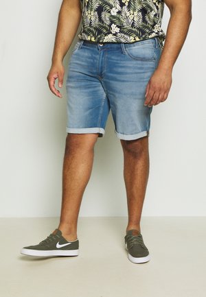 Man in opgerolde lichtblauwe spijkerbroekshorts, groene Nike sneakers met witte swoosh en een zwart bloemenhemd met korte mouwen.
