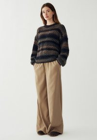 Hravý pruhovaný pletený sveter v tmavomodrej, sivej a béžovej farbe, skombinovaný s béžovými širokými nohavicami a tmavými topánkami so zlatými akcentmi.