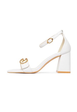 White heeled sandal with block heel, adjustable ankle strap, and gold circular accent on front strap. Smooth texture, minimal design.