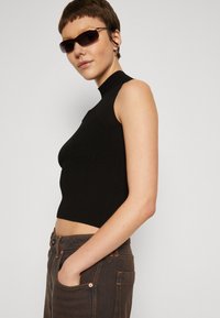 Schwarzes geripptes Crop-Top mit einem Mock-Neck-Design. Das Model trägt eine dunkle Sonnenbrille, kombiniert mit braunen Jeans, die sichtbare Nähte und Taschen aufweisen.