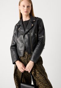 Veste en cuir noir de style motard avec des garnitures en argent, des poches zippées et une coupe courte, associée à un pantalon à imprimé léopard marron.