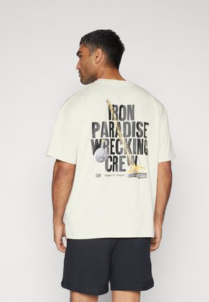 Lyse beige bomuld T-shirt med et stort grafikprint på ryggen med sort tekst og byggematerialer. Korte ærmer og afslappet pasform.