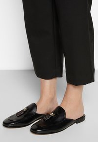 Chaussures slip-on en cuir noir avec un bout rond et un gland décoratif. Texture lisse avec une finition brillante, présentant une semelle à profil bas.