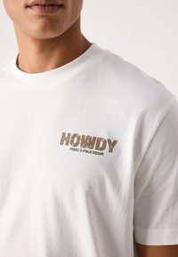 Hvit bomull t-skjorte med en "HOWDY" grafikk i brun og grønn brodering på venstre brystområde. Korte ermer, rund hals.
