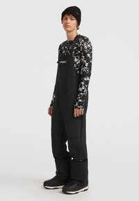 Zwarte waterproof overall met verstelbare banden, voorzak en verstevigde knieën, gecombineerd met een bloemenbedrukt shirt met lange mouwen en een zwarte beanie.