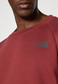 Mørkerød sweatshirt med rund hals, der har et gråt logo fra The North Face på venstre bryst. Tekstureret stof med en blød overflade.
