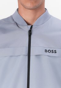 Šviesiai pilkas švarkas su stovinčia apykakle, priekiniais užtrauktukais ir juodu 'BOSS' logotipu. Lygus sintetinis audinys su subtilia tekstūra ir spalvų kontrastu.