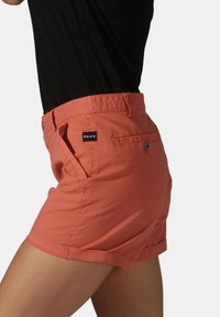 Shorts coral avec une texture lisse, dotés de poches latérales et d'une poche arrière avec fermeture à bouton. Étiquette noire sur la taille.