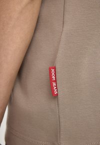 Brązowy bawełniany T-shirt o prążkowanej fakturze, z czerwoną metką wszytą z boku, na której jest napis "JOOP! JEANS." Zbliżenie na boczny detal.