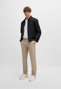 Fekete dzseki, fehér póló, bézs nadrág és fehér sportcipő. A dzsekin cipzár elől és gallér található; a nadrág slim fit fazonú.