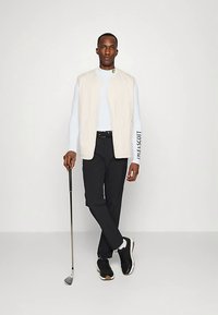 Béžová prošívaná vesta přes světle modrou košili s dlouhým rukávem, doplněná černými kalhotami a černými teniskami. Model drží v ruce golfovou hůl.