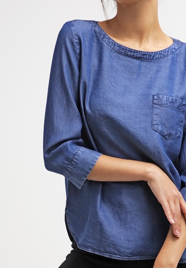 Haut en denim à manches courtes de bleu moyen, col rond, doté d'une poche sur la poitrine gauche et d'un ourlet incurvé. Texture douce avec une coupe décontractée.