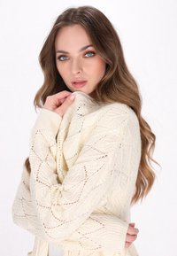 Elfenbeinfarbener Strickcardigan mit Wellenmuster, gerippten Bündchen und offener Vorderseite. Der strukturierte Stoff fällt elegant und verleiht ein weiches, gemütliches Erscheinungsbild.