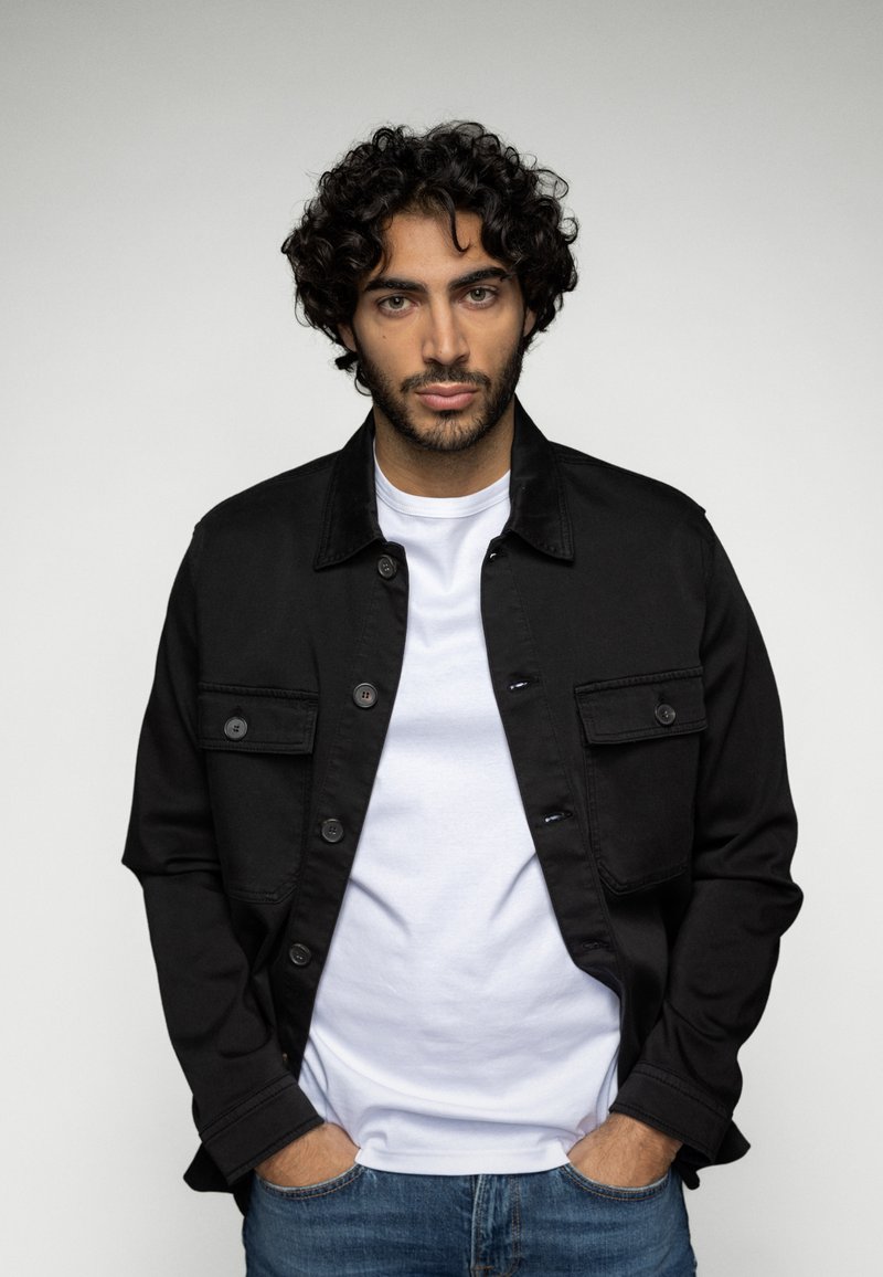 Zwarte spijkerjack met twee voorzakken, gedragen over een witte t-shirt, gecombineerd met blauwe jeans. De stof lijkt glad en stevig.