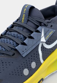 Scarpa da trail Nike con tomaia blu scuro in mesh traspirante e texturizzato, suola gialla, accenti riflettenti e sistema di allacciatura a zigzag.