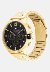 Montre en acier inoxydable doré avec un cadran noir, dotée de trois sous-cadrans, de grands indicateurs d'heure et d'un bracelet au design maillé.