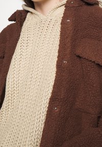 Brun fleecejacka med tryckknappar, lagd över en ljusbeige stickad huva med en kappa och löst, strukturerat stickmönster.