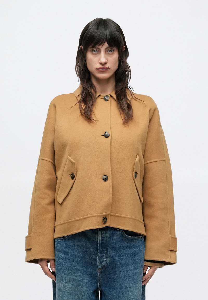 Veste en mélange de laine couleur chameau avec un col, de grandes poches plaquées et des manches oversize. Présente des boutons foncés et un ourlet raccourci.