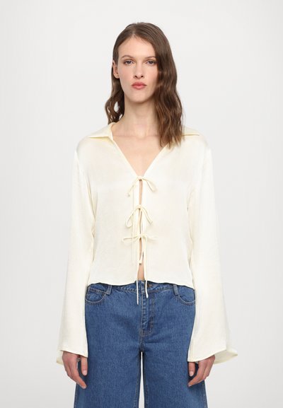Cream-colored blouse with a V-neck and tie-front detail. Features long bell sleeves, textured fabric, and a cropped hem. Paired with blue denim.