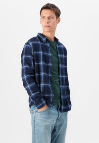 Navy plaid flannel shirt with blue and white check patterns, layered over a dark green t-shirt and paired with light blue denim jeans.