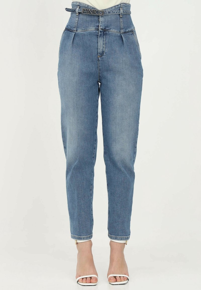 Jean en denim bleu taille haute avec plis, ceinture de marque "PINKO", jambes fuselées, porté avec des escarpins blancs à bout ouvert.