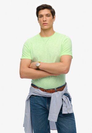 Light green, short-sleeve t-shirt with a subtle texture; paired with blue jeans and a blue striped shirt tied around the waist. Brown belt and watch.