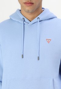 Svetlo modrá mikina s kapucňou vyrobená z bavlneného zmesi, s malým červeným a bielym trojuholníkovým logom na hrudi, nastaviteľnými šnúrkami a prednou kapsou.