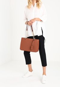 Sac à main en cuir marron avec des poignées en chaîne et en sangle, forme rectangulaire, matière texturée, associé à une chemise blanche et un pantalon noir.