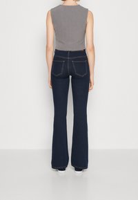 Kvinna som bär en grå ärmlös topp och mörkblå högmidjade utsvängda jeans, stående mot en enkel vit bakgrund.