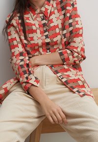 Camicia a maniche lunghe con motivo rosso e beige, con apertura a bottoni e colletto. Indossata con pantaloni a coste di colore crema.